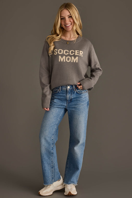 A smiling woman with long blonde hair models the Panache Apparel Collection Soccer Mom Sweater in gray, paired with blue jeans and sneakers, standing against a plain brown background.