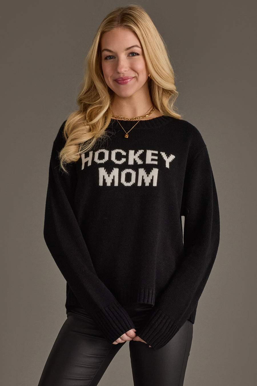 A woman with long blonde hair is wearing the Panache Apparel Collection Black Hockey Mom Sweater, paired with black leather pants and a gold necklace. She stands against a neutral background, smiling softly.
