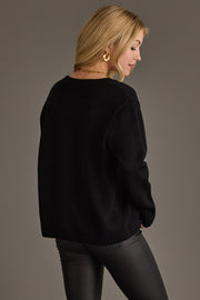 A woman with blonde hair, wearing gold hoop earrings and black pants, stands against a plain gray background, facing away and looking over her shoulder while modeling the Black Hockey Mom Sweater by Panache Apparel Collection.