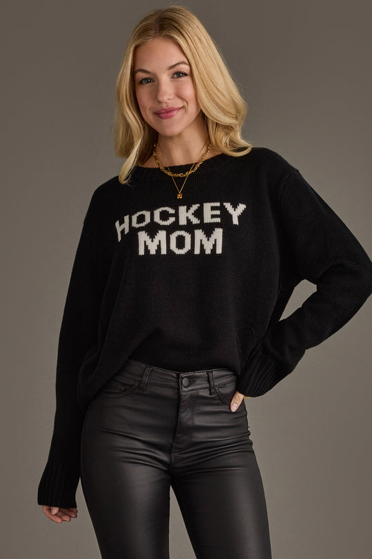 A woman with blonde hair is smiling as she stands against a plain gray background, wearing the Panache Apparel Collection Black Hockey Mom Sweater and black leather pants. The sweater features "HOCKEY MOM" in bold white letters.