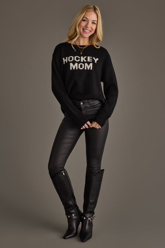 A smiling woman with long blonde hair wears the Panache Apparel Collection Black Hockey Mom Sweater, paired with black leather pants and tall black boots, standing against a plain gray background.