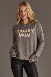 A woman with blonde hair wears the Gray Hockey Mom Sweater from Panache Apparel Collection, featuring beige lettering, along with black pants, standing against a plain gray background.
