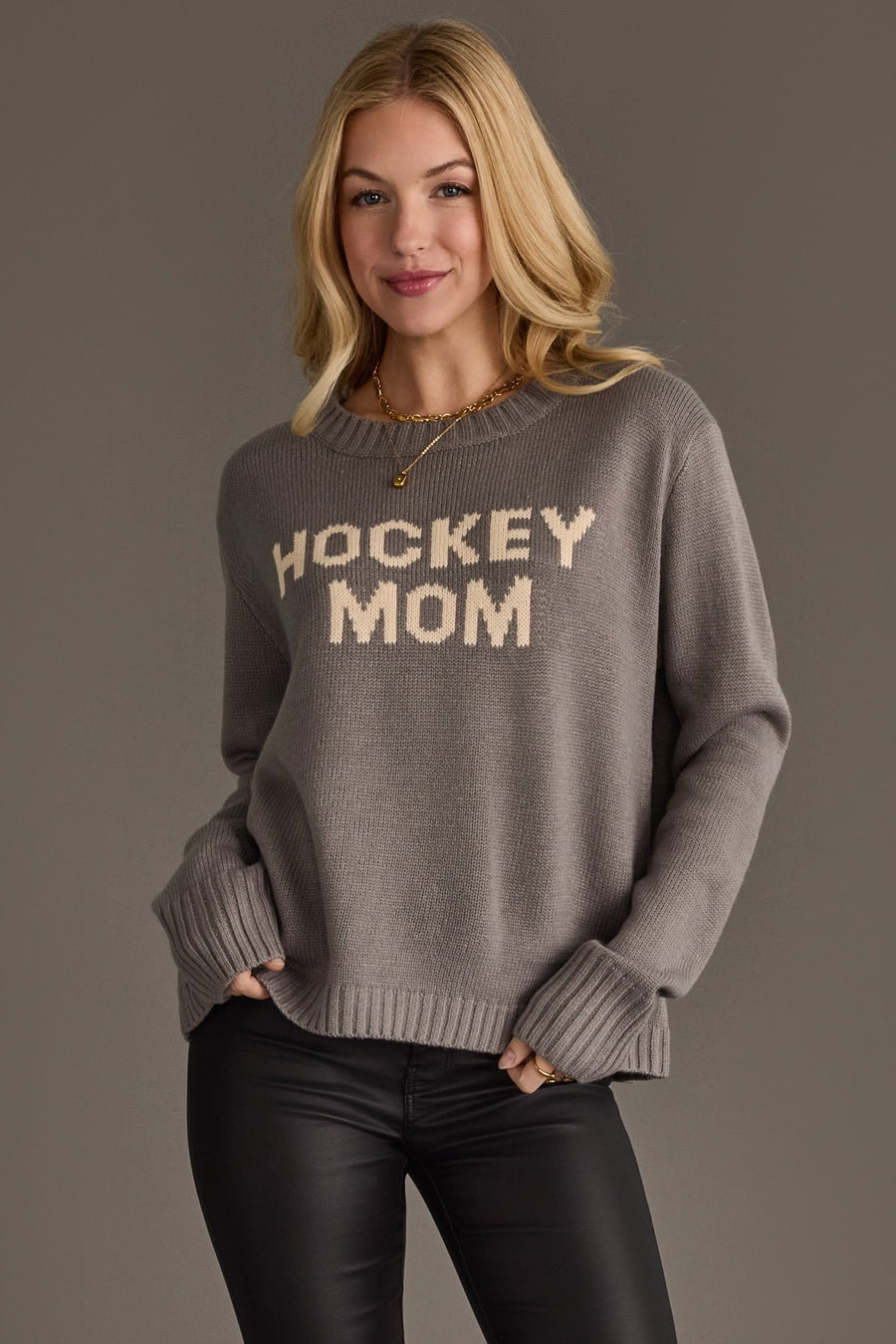A woman with blonde hair wears the Gray Hockey Mom Sweater from Panache Apparel Collection, featuring beige lettering, along with black pants, standing against a plain gray background.