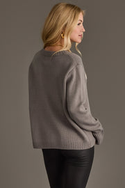 A woman with blonde hair stands facing to the right against a plain grey background, wearing the Gray Hockey Mom Sweater by Panache Apparel Collection and black pants.
