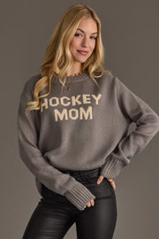 A woman with long blonde hair models the Panache Apparel Collection's Gray Hockey Mom Sweater, featuring bold white lettering, styled with black pants and smiling against a plain gray background.