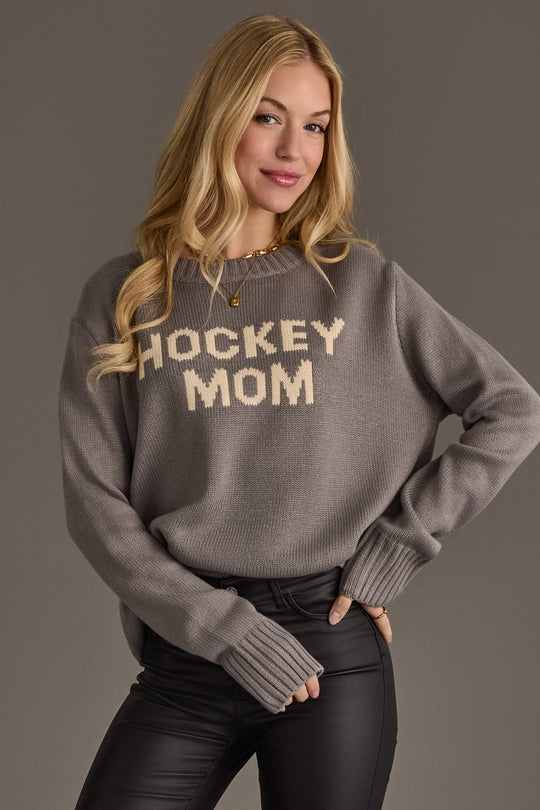 A woman with long blonde hair models the Panache Apparel Collection's Gray Hockey Mom Sweater, featuring bold white lettering, styled with black pants and smiling against a plain gray background.