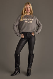 A woman stands against a gray background wearing the Gray Hockey Mom Sweater by Panache Apparel Collection, paired with black leather pants and black boots. She has long blonde hair and exudes confidence with her hands on her hips.