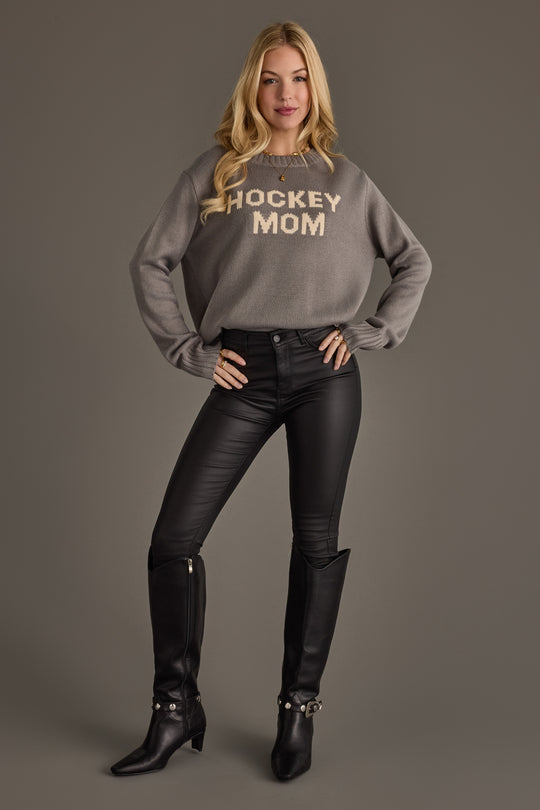 A woman stands against a gray background wearing the Gray Hockey Mom Sweater by Panache Apparel Collection, paired with black leather pants and black boots. She has long blonde hair and exudes confidence with her hands on her hips.