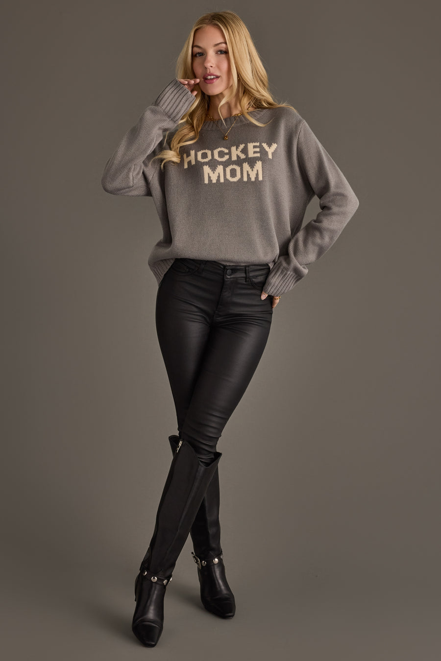 A person with long blonde hair wears the Panache Apparel Collection Gray Hockey Mom Sweater, paired with black pants and black knee-high boots, posing against a plain gray background.
