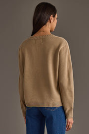 A woman with long dark hair, wearing large hoop earrings, models the Cider Season Sweater by Panache Apparel Collection and blue jeans as she stands facing away from the camera against a plain gray background.