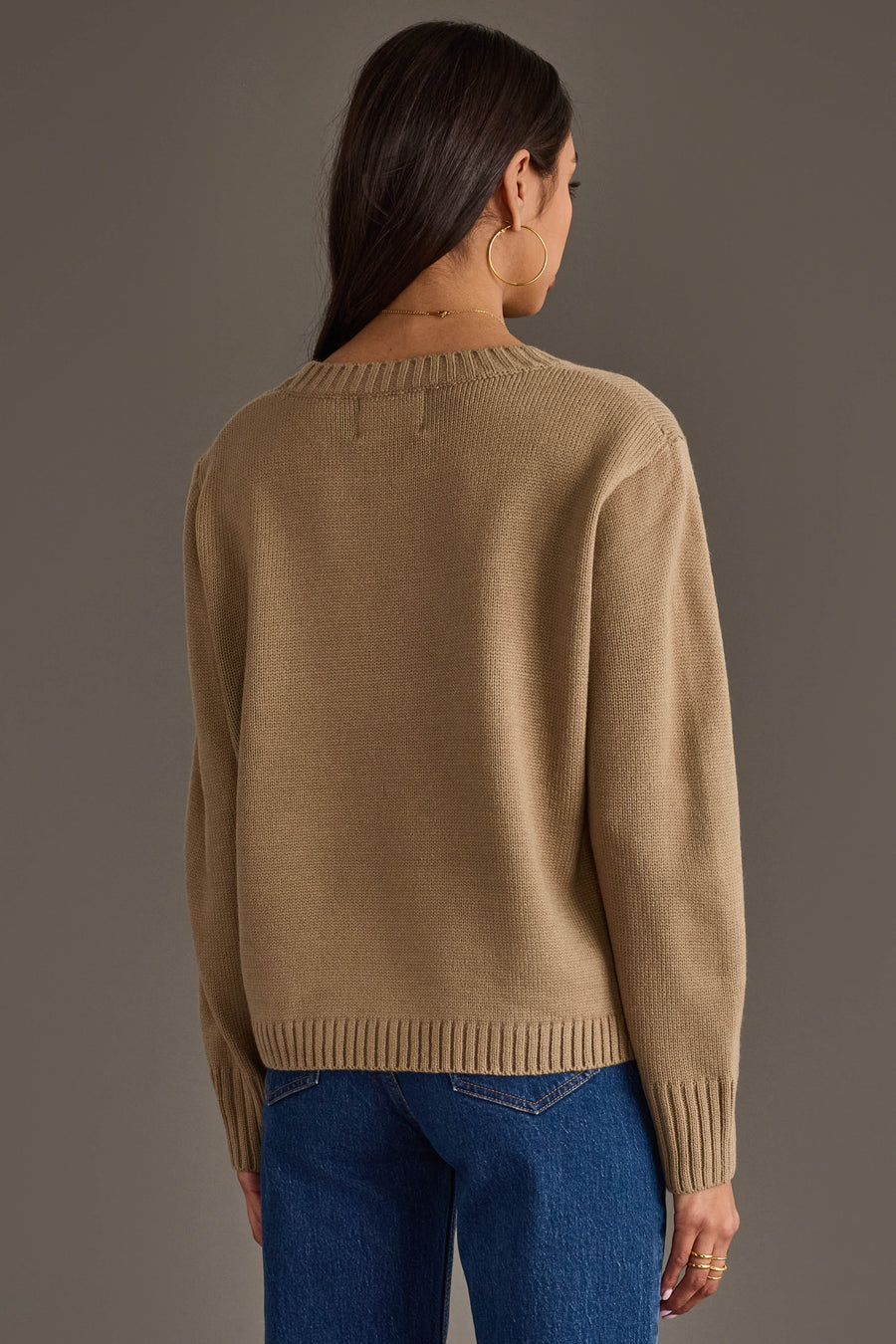 A woman with long dark hair, wearing large hoop earrings, models the Cider Season Sweater by Panache Apparel Collection and blue jeans as she stands facing away from the camera against a plain gray background.