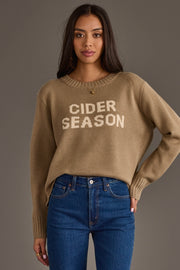 A woman with long dark hair wears the Panache Apparel Collection's Cider Season Sweater, a tan knit featuring “CIDER SEASON” text, paired with blue jeans as she stands against a plain gray background.