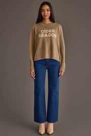 A woman faces forward against a plain gray background, wearing the Panache Apparel Collection's Cider Season Sweater—a tan pullover with "CIDER SEASON" in white lettering—paired with blue jeans and beige boots.
