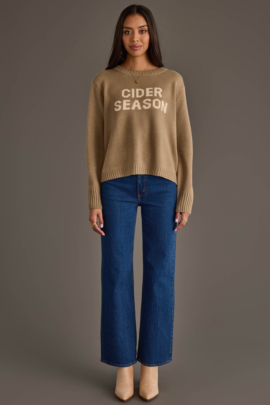 A woman faces forward against a plain gray background, wearing the Panache Apparel Collection's Cider Season Sweater—a tan pullover with "CIDER SEASON" in white lettering—paired with blue jeans and beige boots.