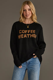 A woman wearing the Black Coffee Weather Sweater from Panache Apparel Collection, featuring "COFFEE WEATHER" in brown letters, and blue jeans stands against a plain gray background, smiling with her hands in her pockets.