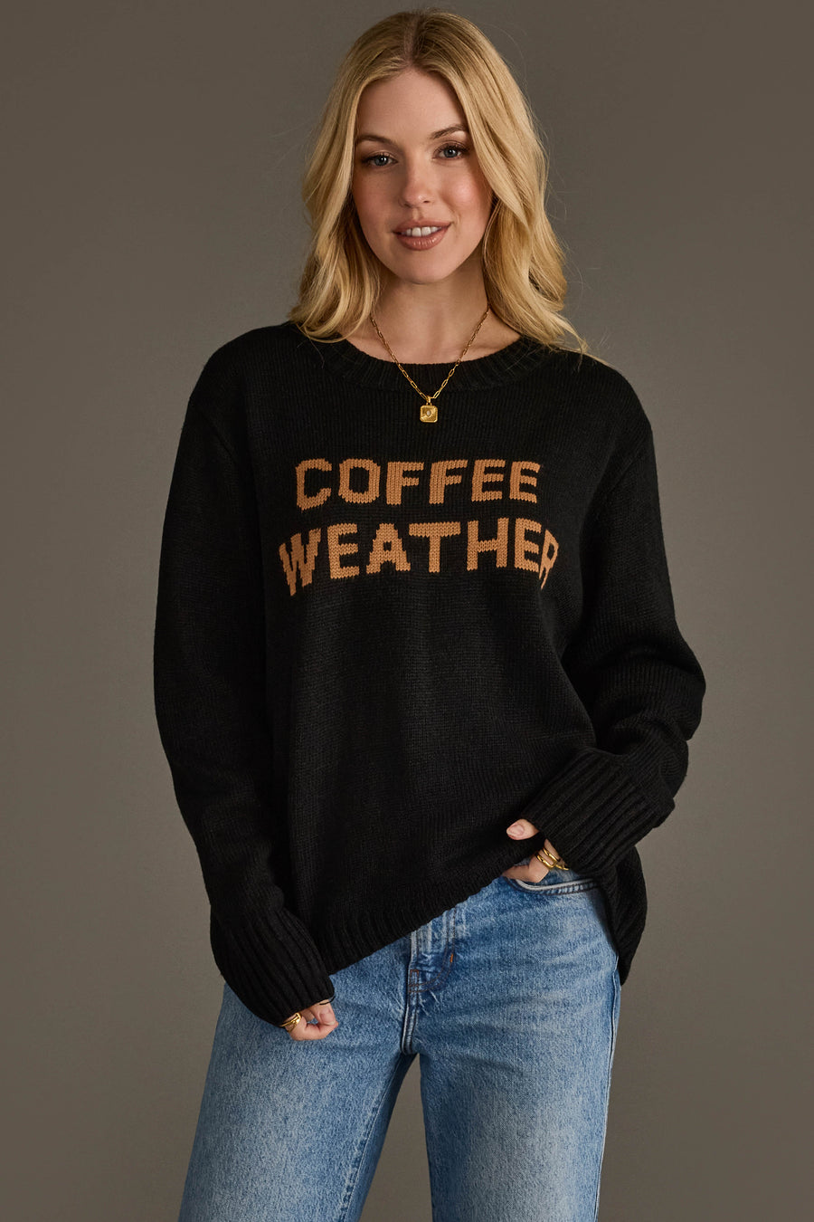 A woman wearing the Black Coffee Weather Sweater from Panache Apparel Collection, featuring "COFFEE WEATHER" in brown letters, and blue jeans stands against a plain gray background, smiling with her hands in her pockets.