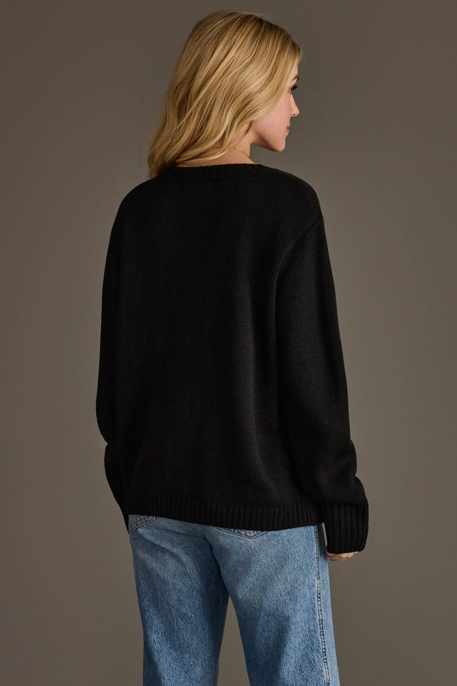 A person with blonde hair, seen from behind, wearing the Black Coffee Weather Sweater by Panache Apparel Collection and blue jeans, stands in front of a plain brown background.