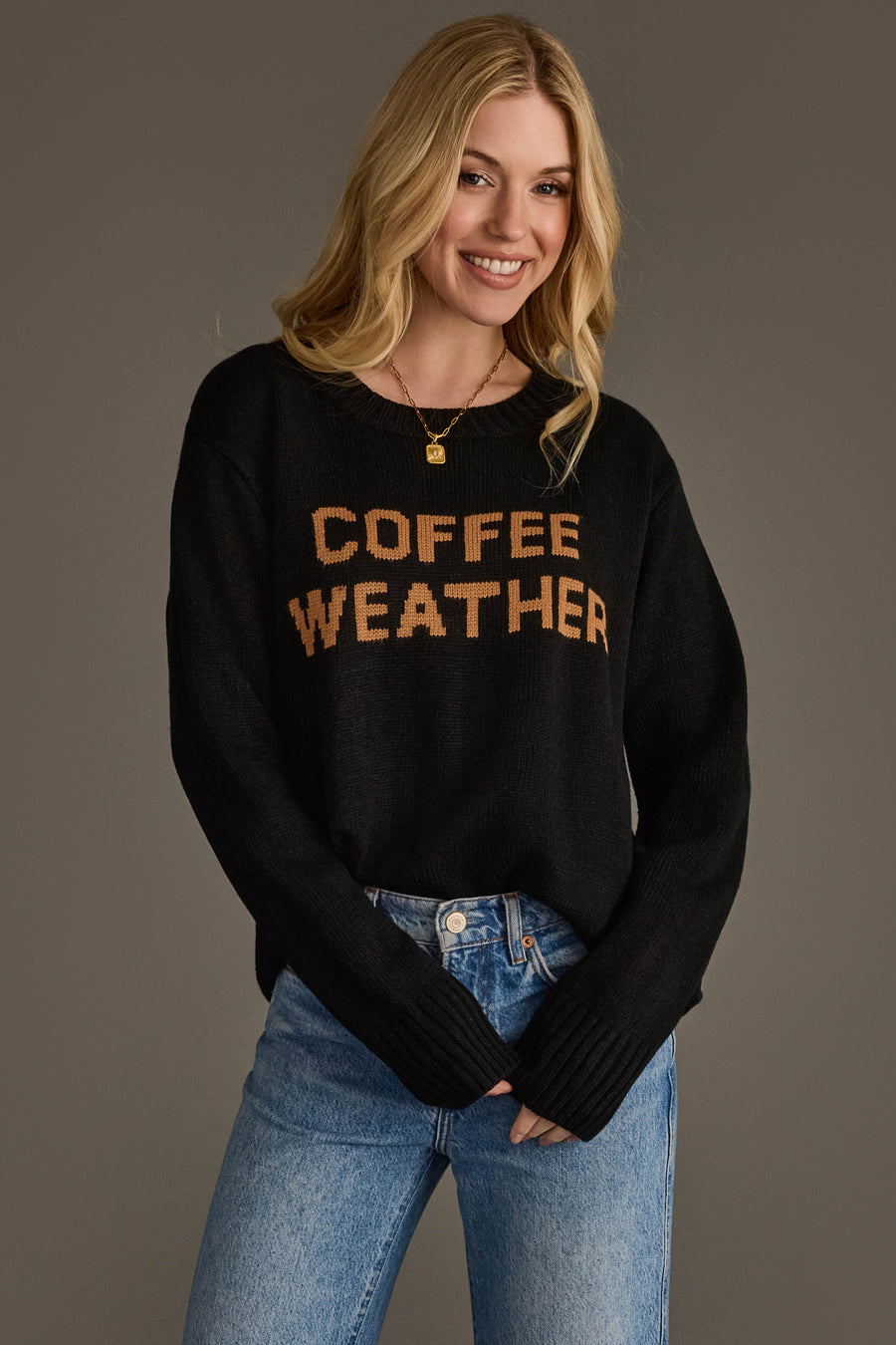 A woman stands smiling against a plain background, wearing the Panache Apparel Collection Black Coffee Weather Sweater paired with blue jeans.