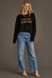 A woman stands against a gray background, smiling slightly as she models the Panache Apparel Collection's Black Coffee Weather Sweater featuring bold orange lettering. She pairs it with light blue jeans and beige heels, her long blonde hair flowing loosely.