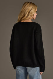 A person with long blonde hair, wearing the Panache Apparel Collection’s Black & Tan Whiskey Weather Sweater and light blue jeans, is standing with their back to the camera against a plain, dark gray background.