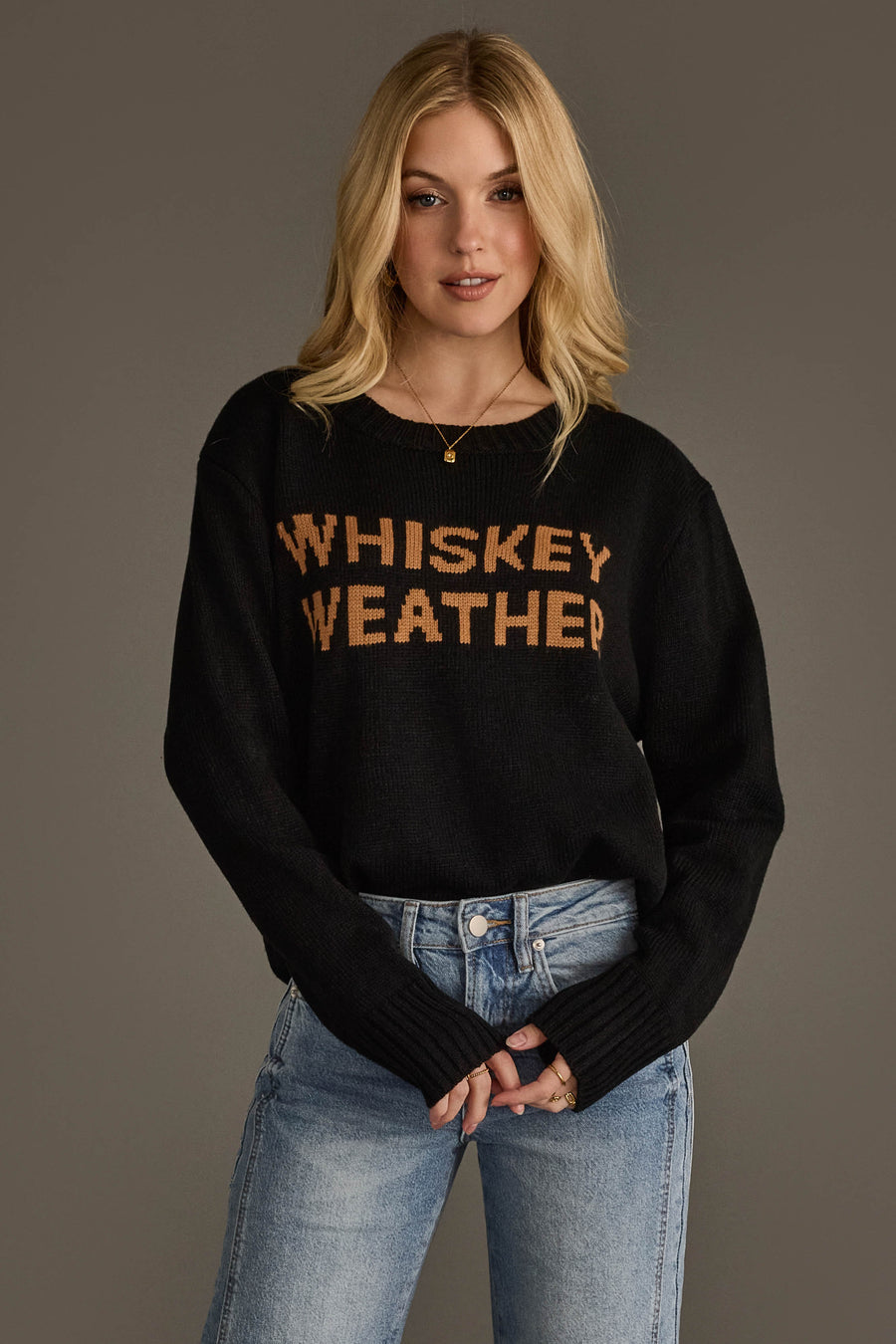 A woman with blonde, loosely waved hair wears the Panache Apparel Collection's Black & Tan Whiskey Weather Sweater, featuring “WHISKEY WEATHER” in brown lettering, paired with light blue jeans and set against a plain gray background.