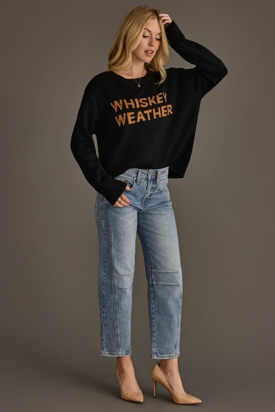A woman with blonde hair stands against a gray background, wearing the Panache Apparel Collection Black & Tan Whiskey Weather Sweater, paired with light blue jeans and tan high heels. She is touching her head with one hand.
