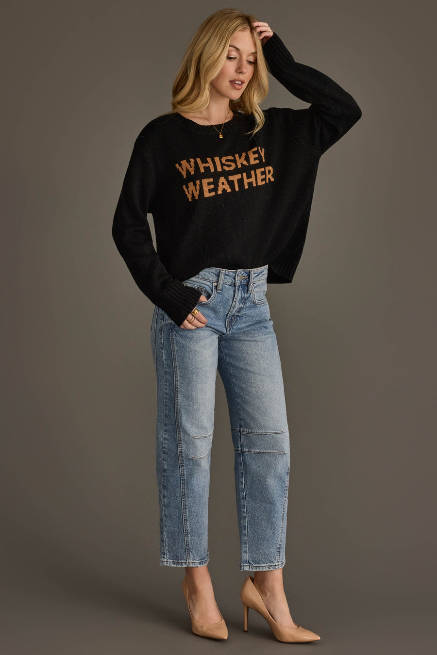 A woman with blonde hair stands against a gray background, wearing the Panache Apparel Collection Black & Tan Whiskey Weather Sweater, paired with light blue jeans and tan high heels. She is touching her head with one hand.