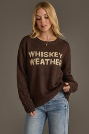A woman wearing the Panache Apparel Collection’s Dark Brown Whiskey Weather Sweater stands against a gray background, looking down and holding the hem of her sweater with one hand. She is also wearing light blue jeans.