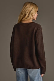 A person with long blonde hair, dressed in the Panache Apparel Collection's Dark Brown Whiskey Weather Sweater and light blue jeans, stands facing away from the camera against a plain gray background.