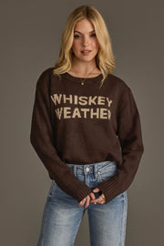 A woman with blonde hair wears the Panache Apparel Collection's Dark Brown Whiskey Weather Sweater and blue jeans, standing against a plain gray background.