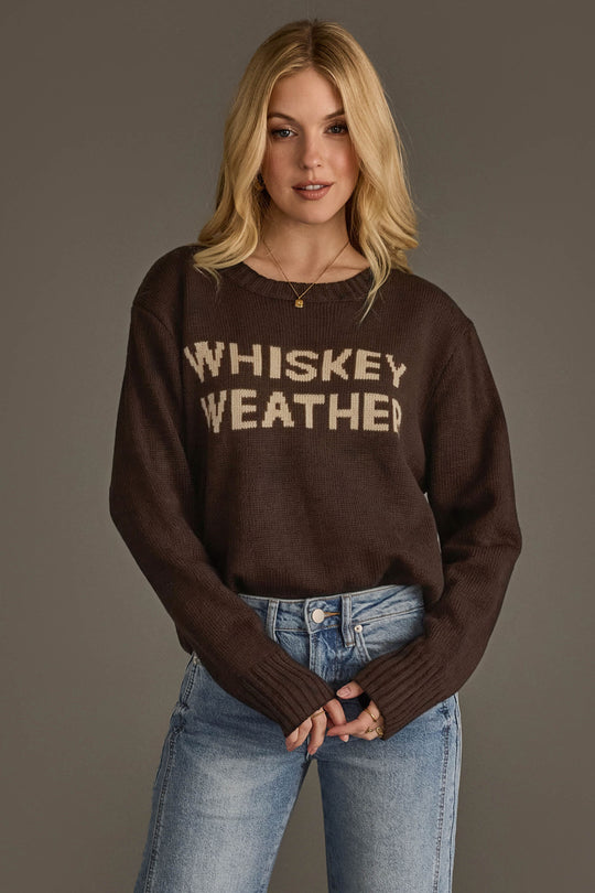 A woman with blonde hair wears the Panache Apparel Collection's Dark Brown Whiskey Weather Sweater and blue jeans, standing against a plain gray background.