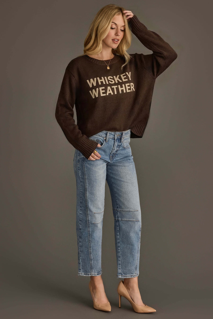 A woman wearing a dark brown Whiskey Weather Sweater from Panache Apparel Collection, paired with light blue jeans and nude heels, stands against a gray background with one hand touching her hair and the other in her pocket.