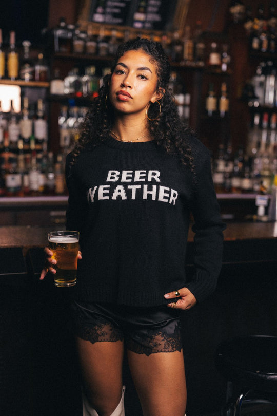 A woman with curly hair stands in a bar holding a glass of beer, wearing the Panache Apparel Collection’s relaxed fit Beer Weather Sweater and black lace-trimmed shorts. Bottles line the shelves in the background.