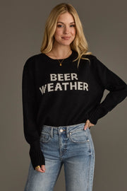 A woman models the Panache Apparel Collection Beer Weather Sweater. A black top featuring "BEER WEATHER" in bold white letters as she smiles with one hand on her hip, paired with blue jeans against a plain background.
