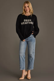 A woman with blonde hair stands against a plain background wearing the Panache Apparel Collection’s Beer Weather Sweater, paired with light blue jeans and beige heels. One hand rests in her pocket.