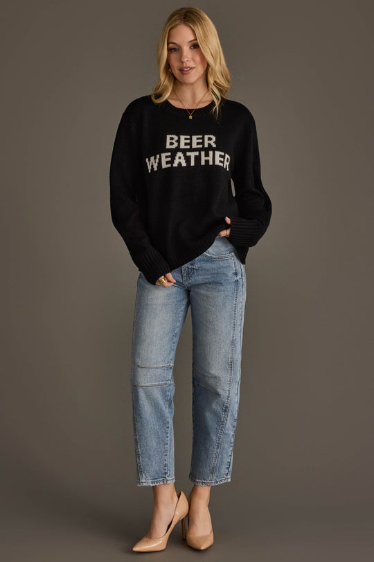 A woman with blonde hair stands against a plain background wearing the Panache Apparel Collection’s Beer Weather Sweater, paired with light blue jeans and beige heels. One hand rests in her pocket.