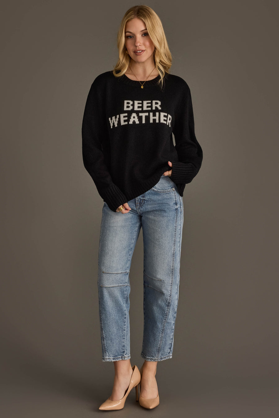 A woman with blonde hair stands against a plain background wearing the Panache Apparel Collection’s Beer Weather Sweater, paired with light blue jeans and beige heels. One hand rests in her pocket.