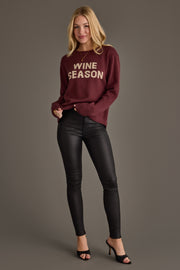 A woman with long blonde hair, dressed in the Panache Apparel Collection Burgundy & Ivory Wine Season Sweater, black fitted pants, and black heeled sandals, stands smiling against a plain gray background.