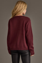 A woman with long blonde hair, seen from behind, wears the Panache Apparel Collection’s Burgundy & Ivory Wine Season Sweater paired with black leather pants, standing against a plain brown background.