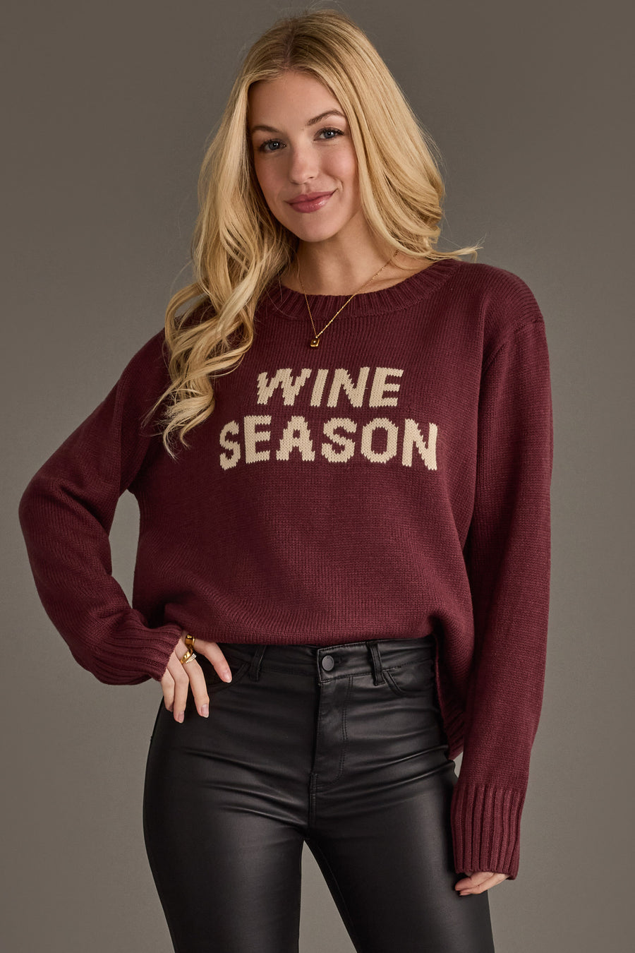 A woman with long blonde hair stands against a neutral background, smiling slightly as she wears the Panache Apparel Collection Burgundy & Ivory Wine Season Sweater paired with black leather pants.