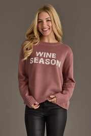 A smiling woman with long blonde hair models the Panache Apparel Collection’s Mauve & White Wine Season Sweater, paired with black pants, against a plain gray background.