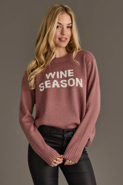 A woman with long blonde hair is standing against a plain gray background, softly smiling as she wears the Panache Apparel Collection Mauve & White Wine Season Sweater and black pants.