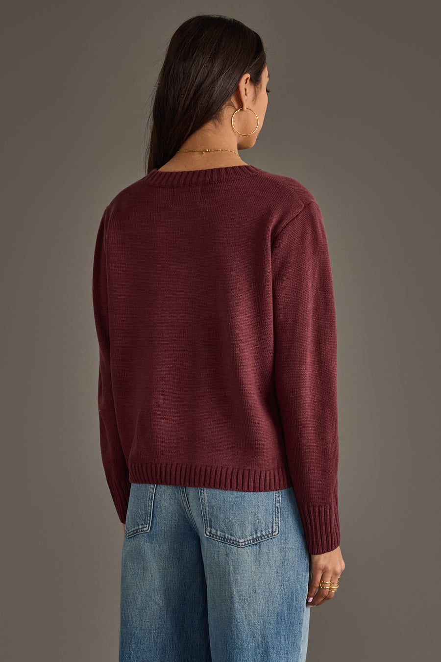 A woman with long brown hair models the Panache Apparel Collection's Burgundy & Light Pink Wine Season Sweater, paired with light blue jeans, as she stands facing away from the camera against a plain brown background.