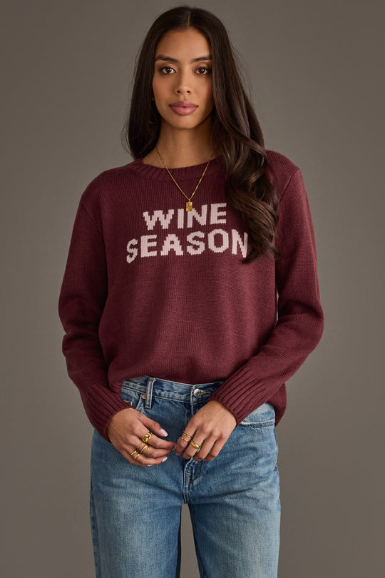 A woman with long dark hair is wearing the Panache Apparel Collection Burgundy & Light Pink Wine Season Sweater and blue jeans, standing against a gray background.