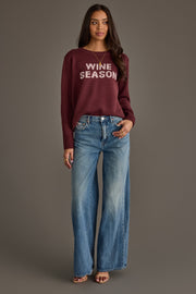 A woman stands facing forward against a plain gray background, wearing the Panache Apparel Collection Burgundy & Light Pink Wine Season Sweater paired with blue wide-leg jeans and heels, her expression relaxed.