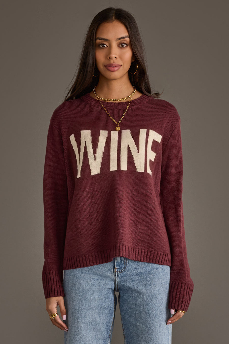 A woman with long dark hair wears the Panache Apparel Collection Burgundy & Ivory Wine Sweater, featuring "WINE" in bold ivory letters, styled with light blue jeans and layered gold necklaces, standing against a plain gray background.