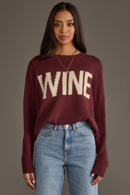 A woman wearing the Panache Apparel Collection Burgundy & Ivory Wine Sweater stands against a plain gray background. She has long dark hair, pairs the sweater with blue jeans, and accessorizes with gold jewelry.