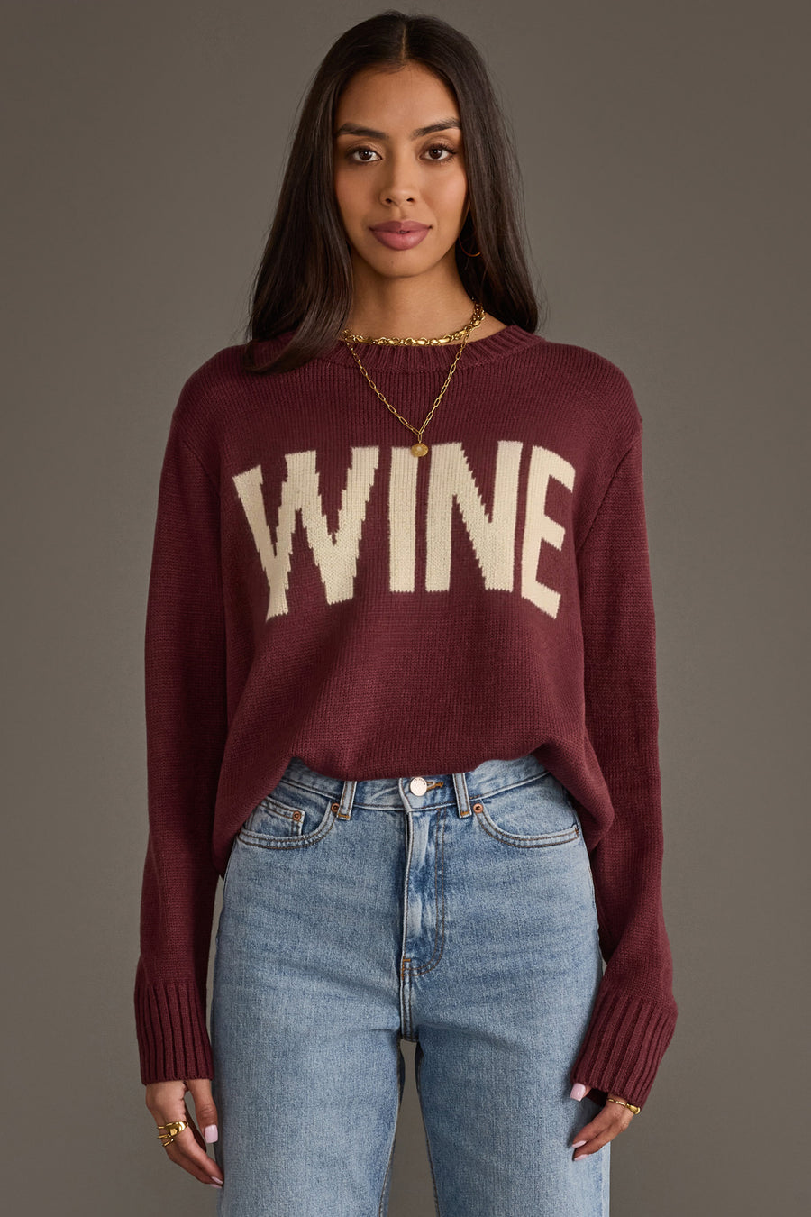 A woman wearing the Panache Apparel Collection Burgundy & Ivory Wine Sweater stands against a plain gray background. She has long dark hair, pairs the sweater with blue jeans, and accessorizes with gold jewelry.