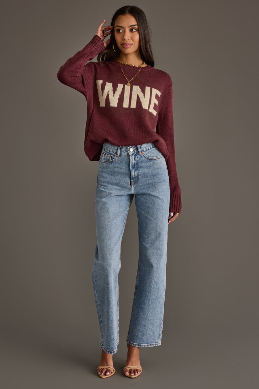 A woman with long dark hair poses against a gray background, wearing the Panache Apparel Collection Burgundy & Ivory Wine Sweater, light-wash blue jeans, and nude heeled sandals. She has her right hand to her head.
