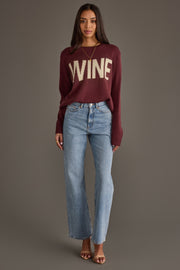 A woman stands against a gray background wearing the Panache Apparel Collection Burgundy & Ivory Wine Sweater, light blue jeans, brown heeled sandals, and a gold necklace.